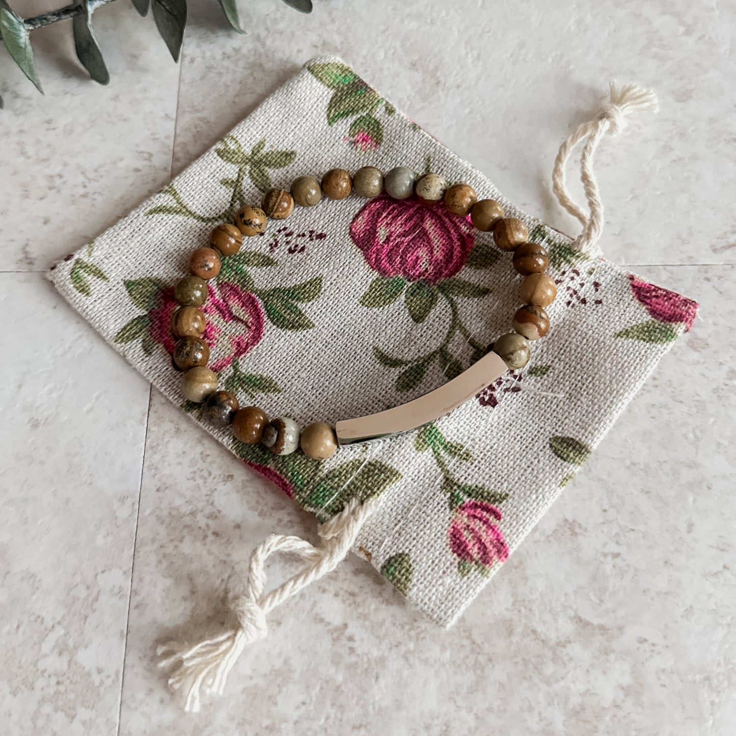 Earthy jasper beaded bracelet with curved stainless steel bar, resting on a floral drawstring pouch.