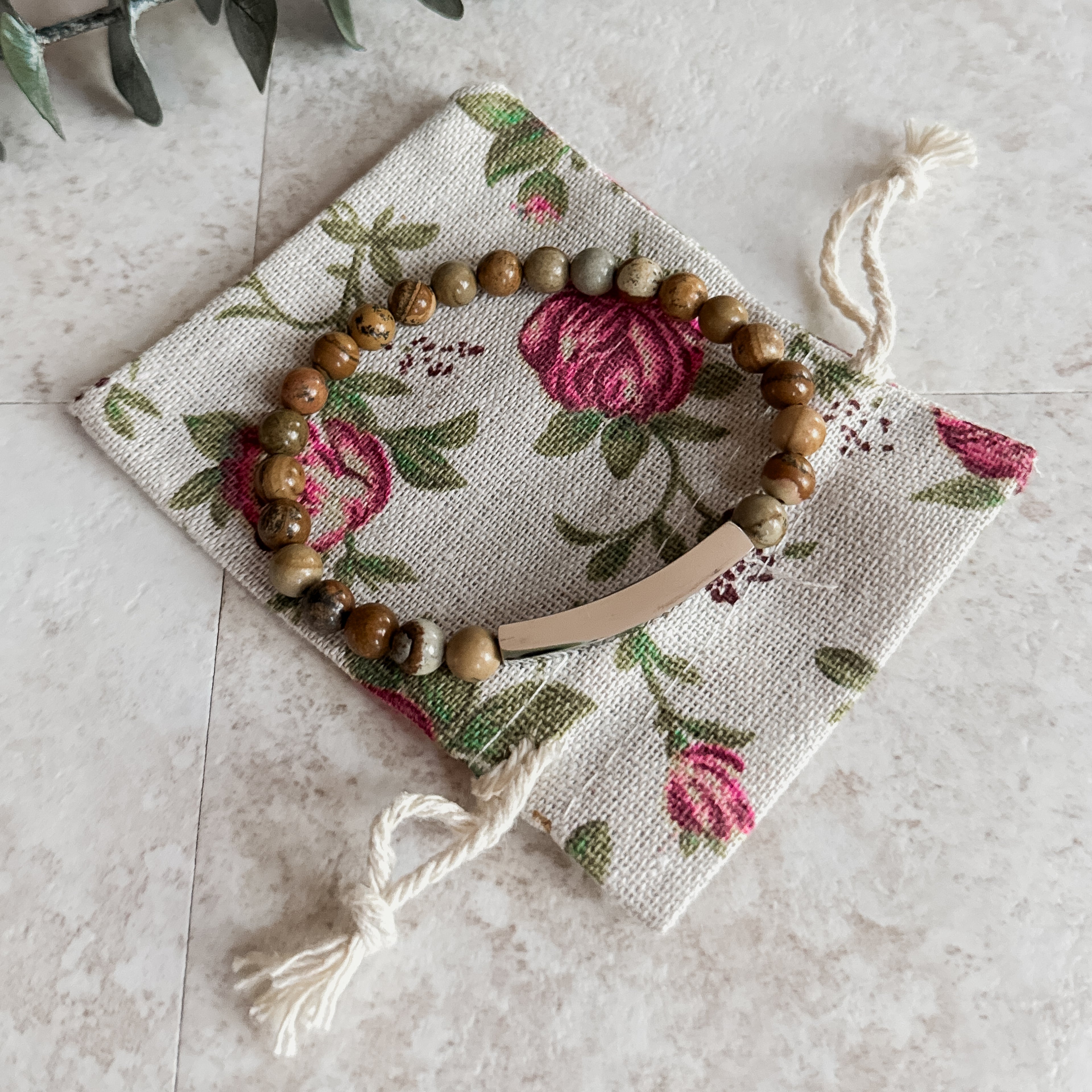 Earthy jasper beaded bracelet with curved stainless steel bar, resting on a floral drawstring pouch.