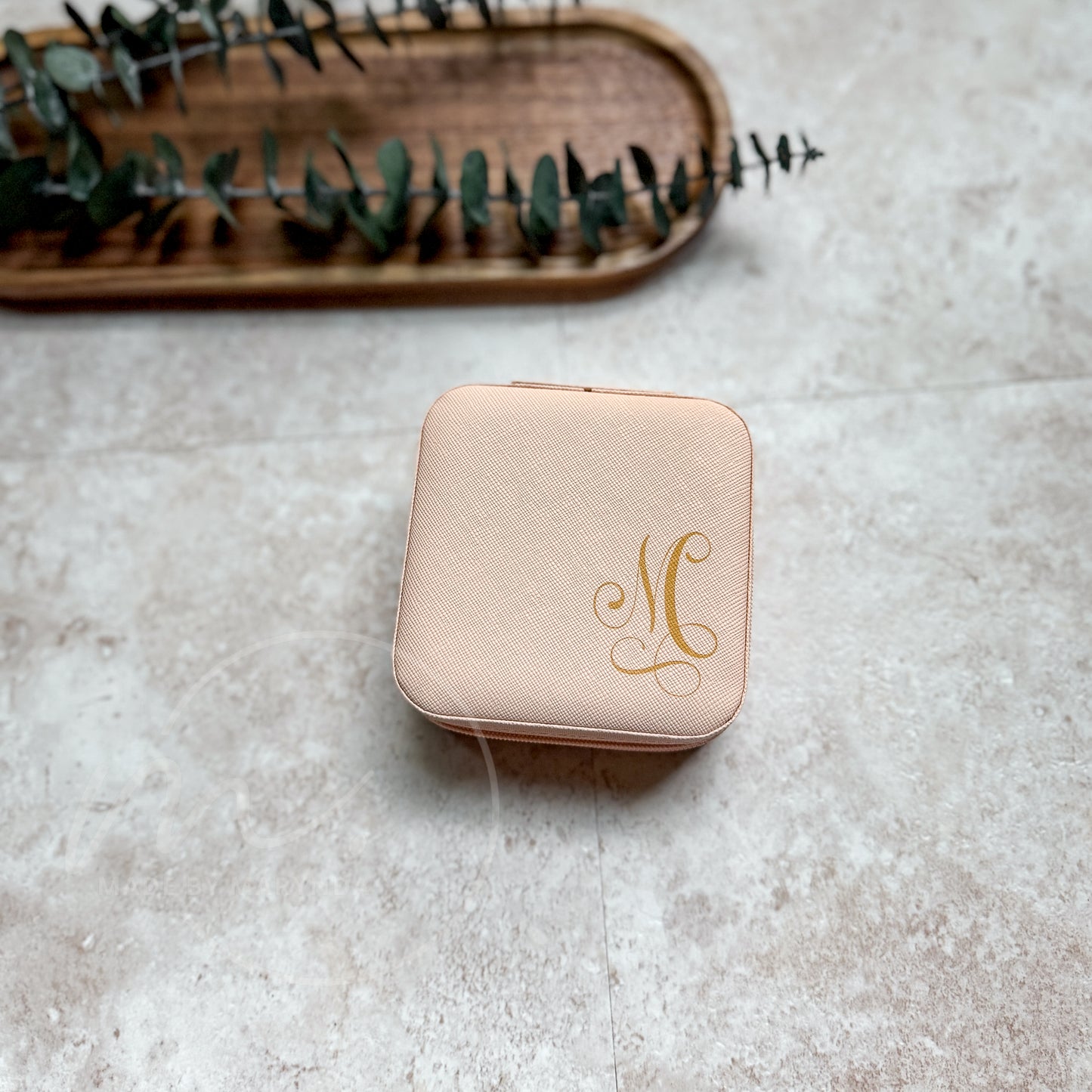 Pink jewelry box with gold monogram on a textured surface with a wooden tray and eucalyptus leaves in the background.