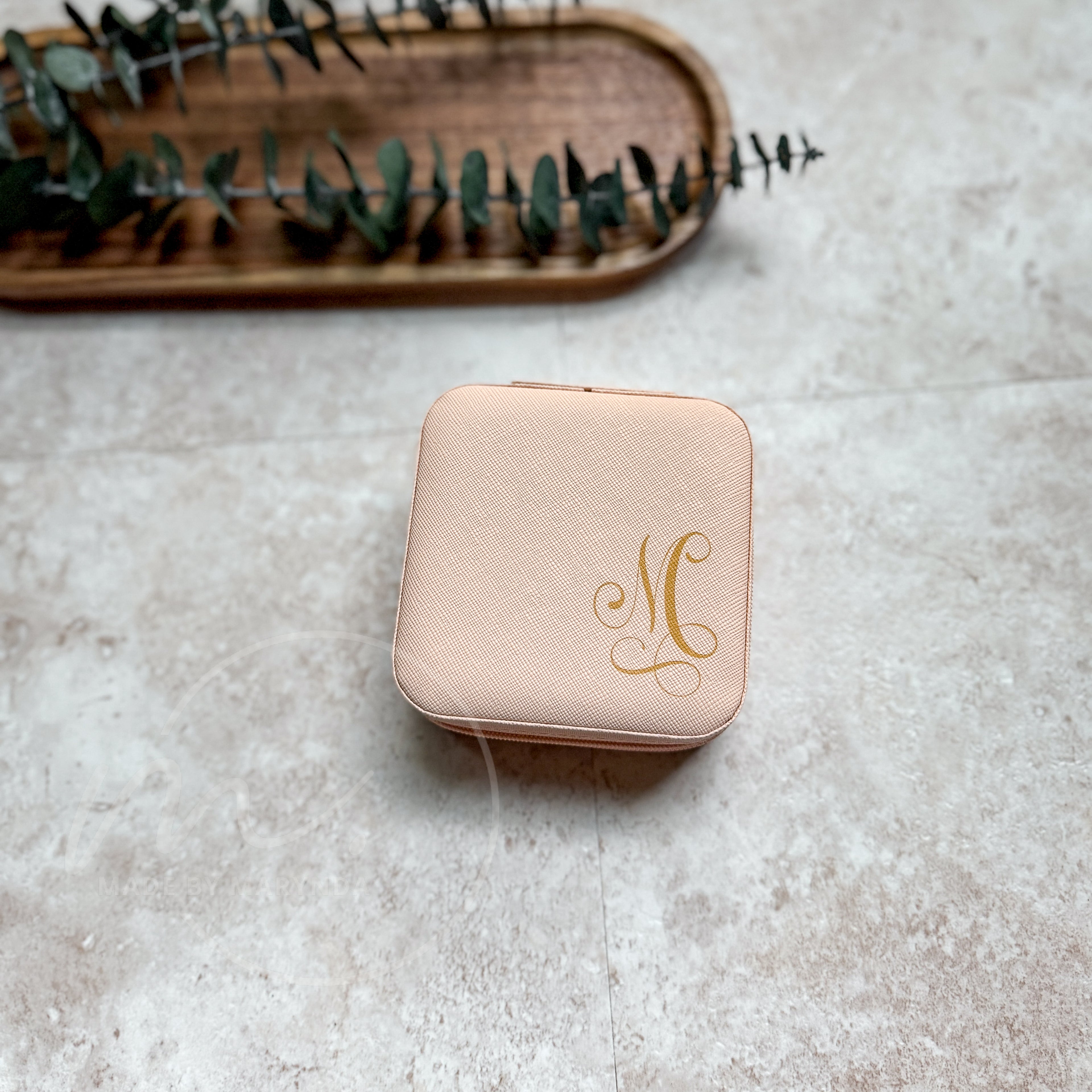Pink jewelry box with gold monogram on a textured surface with a wooden tray and eucalyptus leaves in the background.
