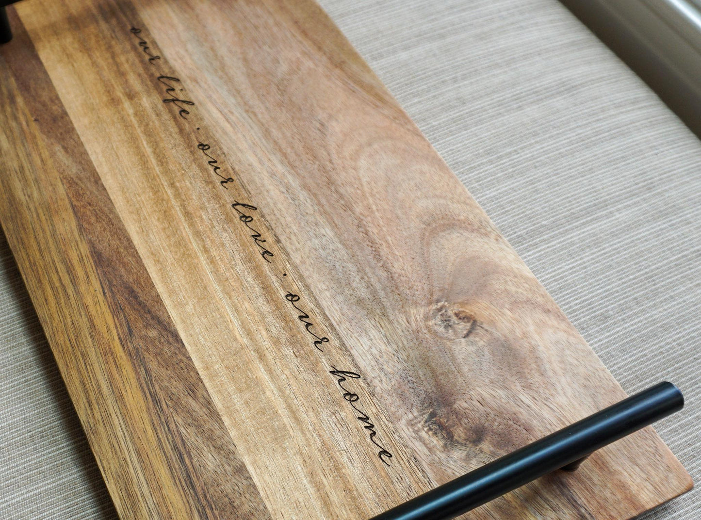 Angled view of acacia wood tray with black handle and engraved quote 'our life · our love · our home' on a linen-textured background