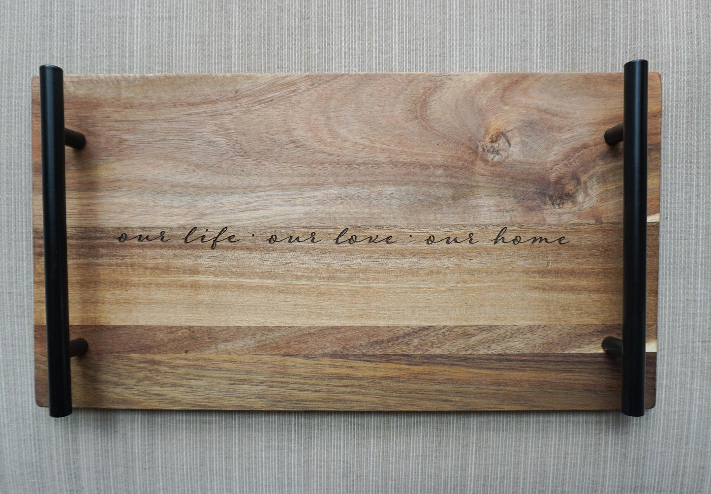 Top-down view of engraved acacia wood tray with black handles and the quote ‘our life · our love · our home’ on a textured gray background