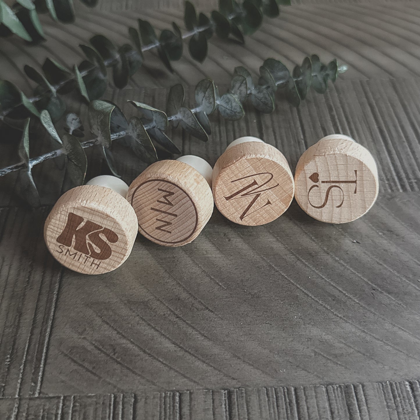 Angled view of four personalized wooden wine corks with engraved initials, shown on a dark wood surface with eucalyptus in the background.