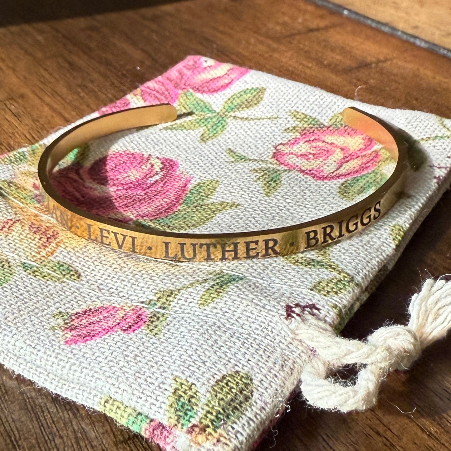 Gold engraved cuff bracelet with names displayed on a floral drawstring pouch over a wooden surface