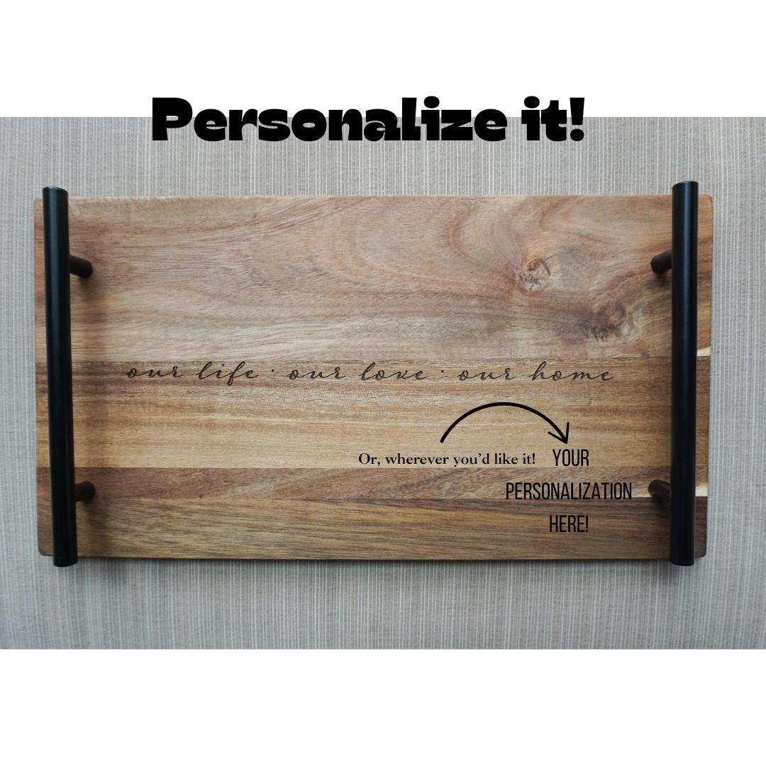 Wooden tray with black handles and engraved quote ‘our life · our love · our home,’ shown with customization example and arrow pointing to personalization area