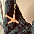 Wooden airplane-shaped keychain with “Home is where my pilot lands” text, attached to a black quilted jacket on a beige background.