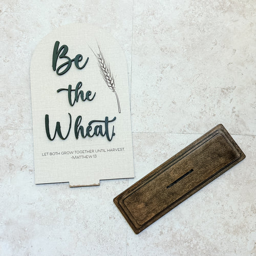 Decorative sign with 'Be the Wheat' text and a wooden tray on a textured surface