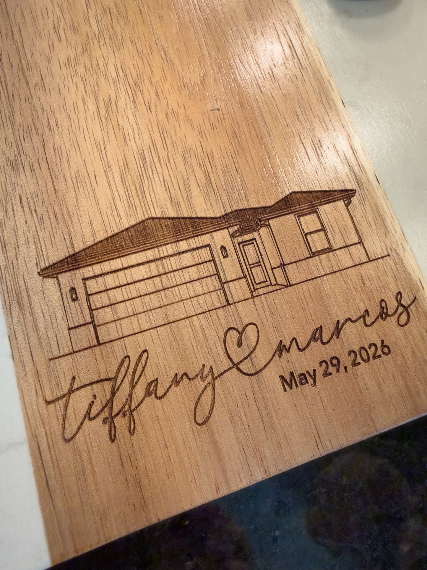 Wooden plaque with engraved house illustration, names 'Tiffany & Marcos', and date 'May 29, 2026'.
