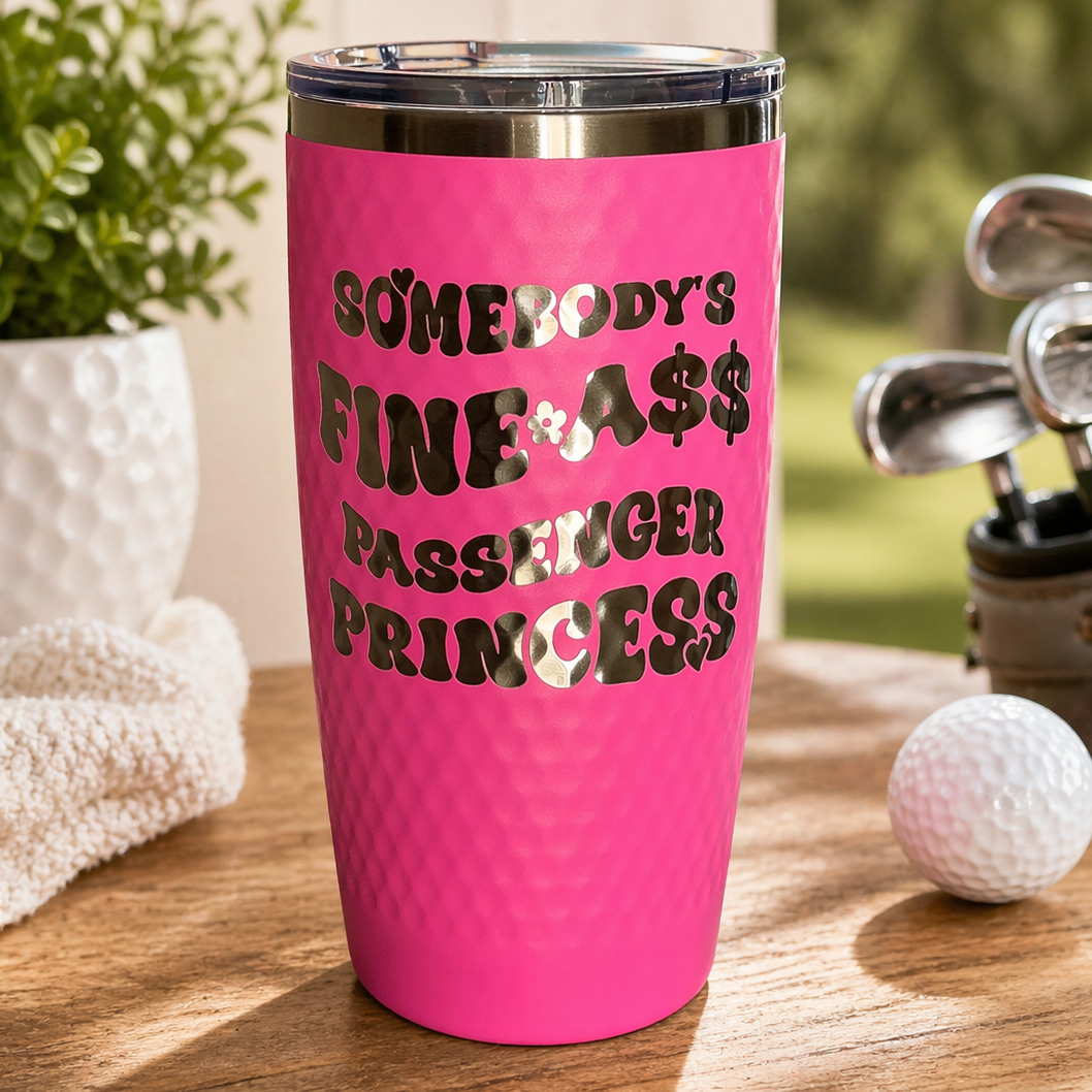 Pink tumbler with text on a table with golf equipment and plants.