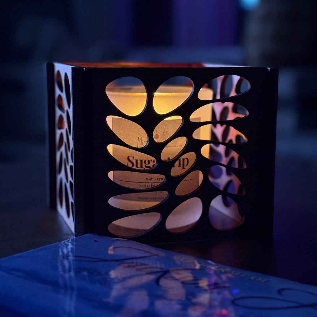 Wooden candle lantern with leaf cut-out design, glowing warmly in a dark room with visible candle branding inside