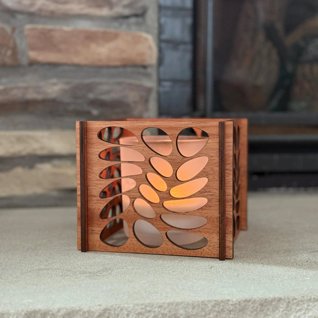 Laser-cut wooden lantern with leaf design glowing from a tealight, placed on a stone surface in front of a rustic fireplace