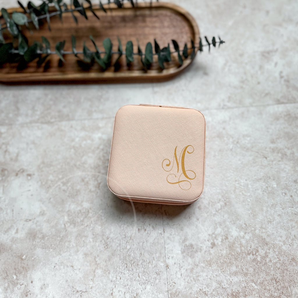 Pink jewelry box with gold monogram on a textured surface with a wooden tray and eucalyptus leaves in the background.