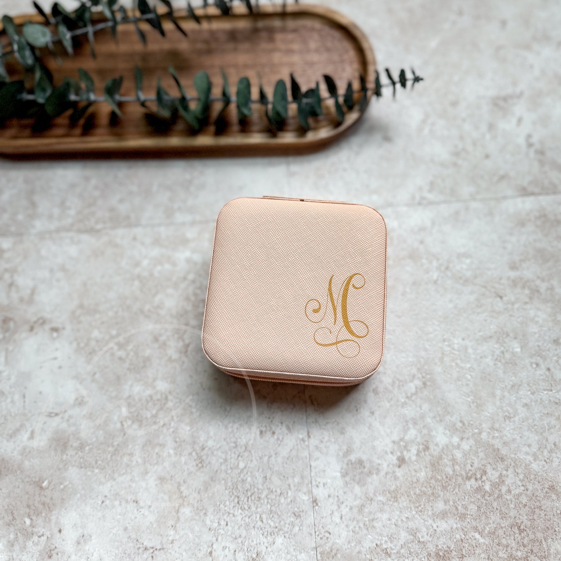 Pink jewelry box with gold monogram on a textured surface with a wooden tray and eucalyptus leaves in the background.