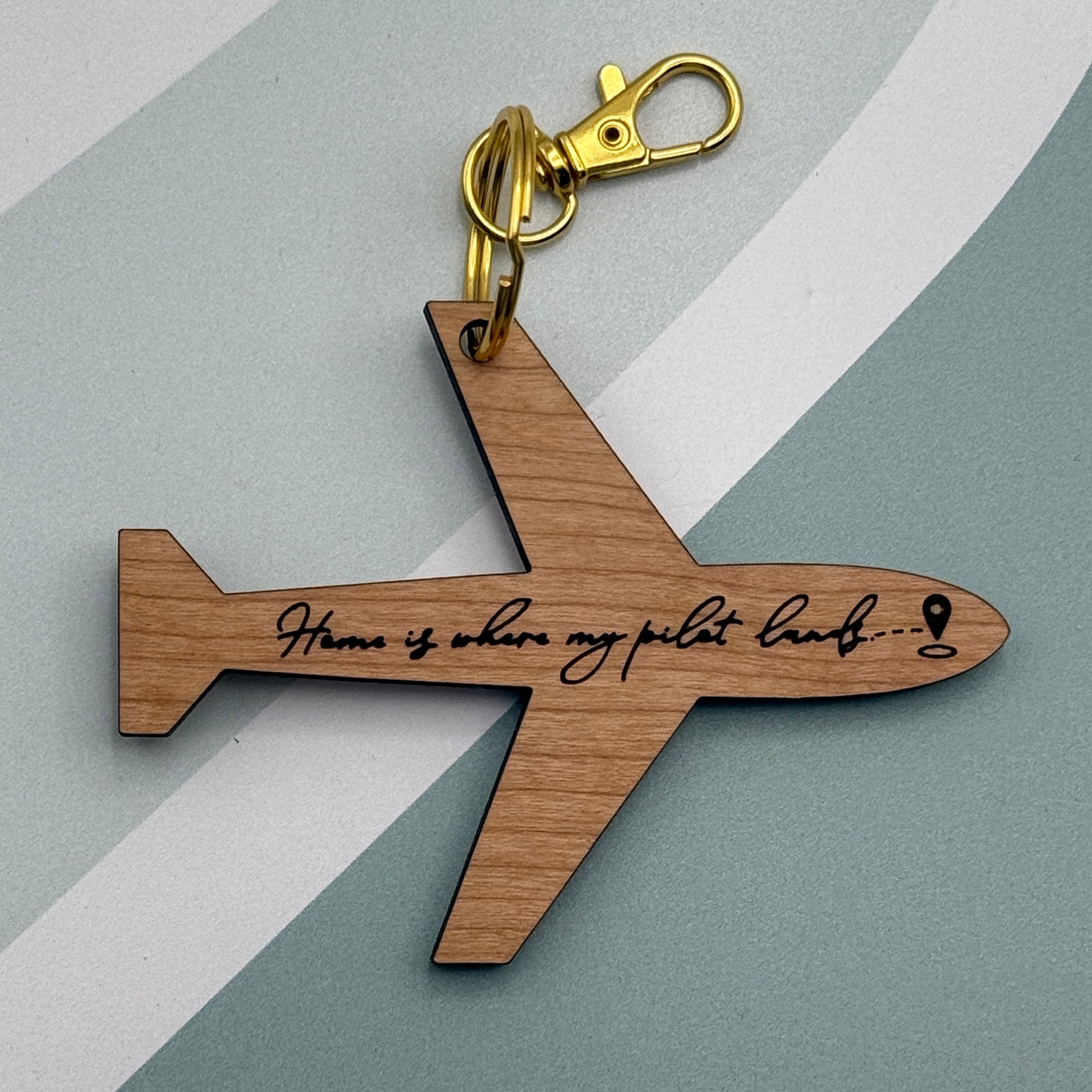 Laser-engraved airplane-shaped keychain with “Home is where my pilot lands” text, shown on a soft gray and white background with gold hardware.