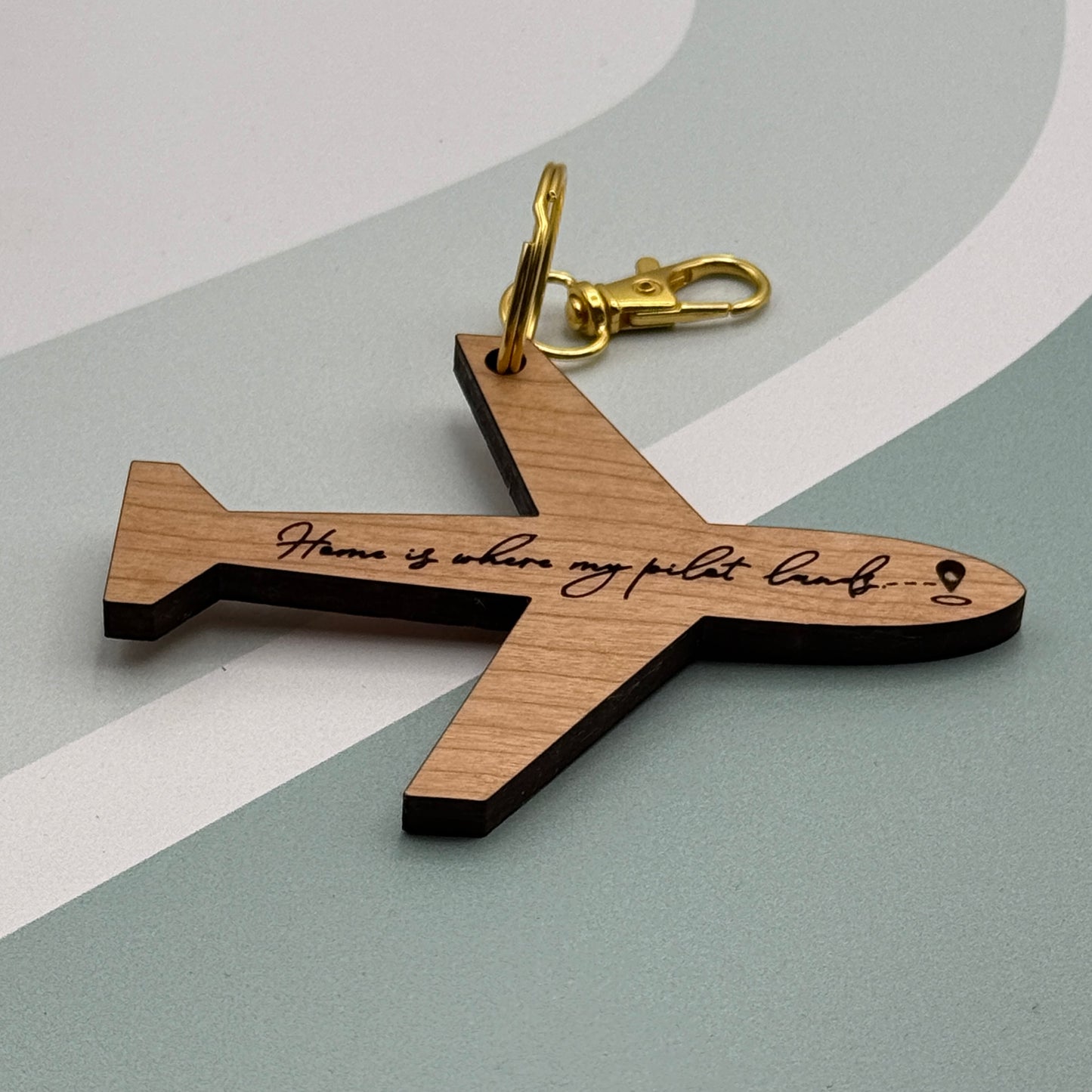 Laser-engraved airplane-shaped keychain with “Home is where my pilot lands” text, shown on a soft gray and white background with gold hardware.