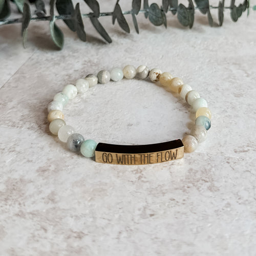 Amazonite bead bracelet with engraved gold bar reading “Go With the Flow,” displayed on a light stone surface.