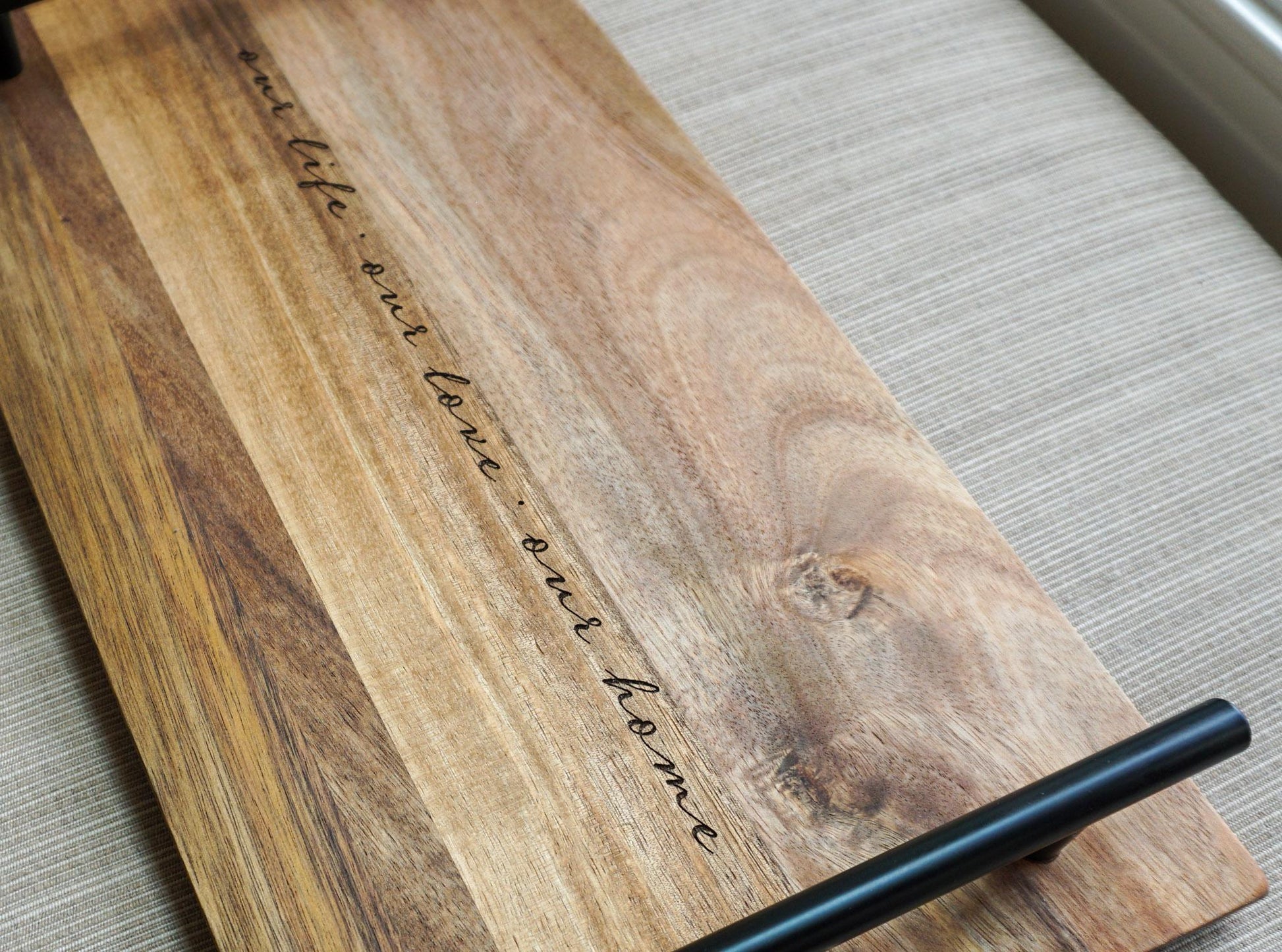 Angled view of acacia wood tray with black handle and engraved quote 'our life · our love · our home' on a linen-textured background