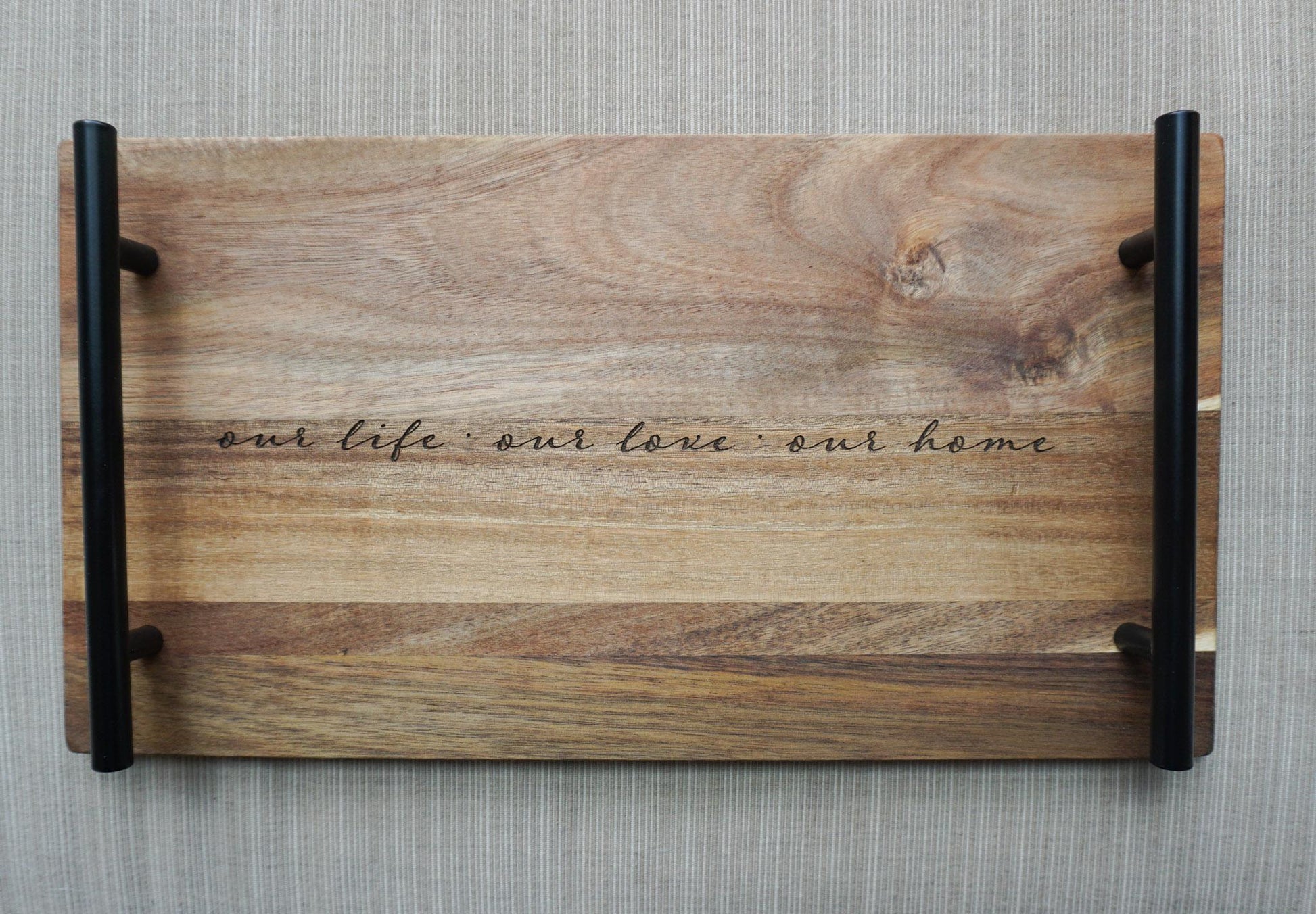 Top-down view of engraved acacia wood tray with black handles and the quote ‘our life · our love · our home’ on a textured gray background