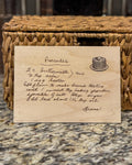 Engraved wooden plaque featuring a handwritten pancake recipe, displayed on a marble counter in front of a woven basket.
