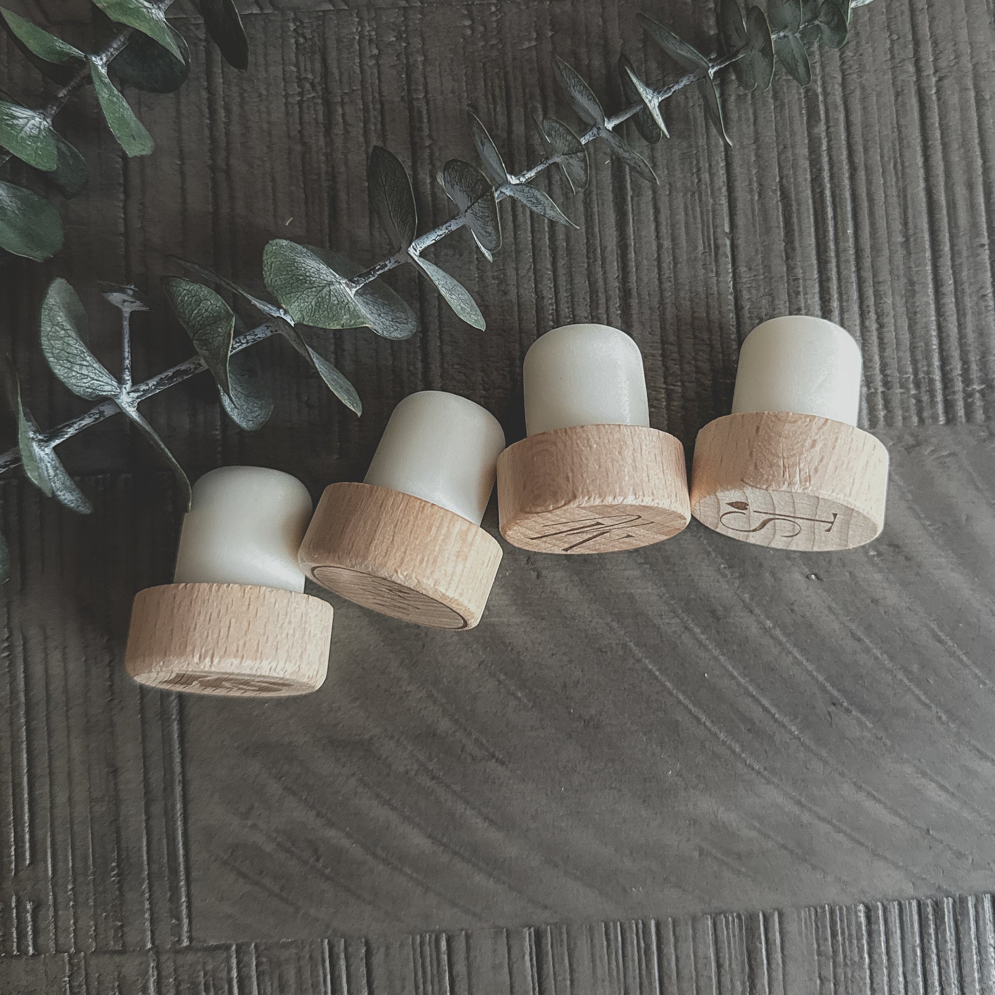Side view of four engraved wooden wine corks with silicone stoppers, displayed on a dark textured surface with eucalyptus greenery.