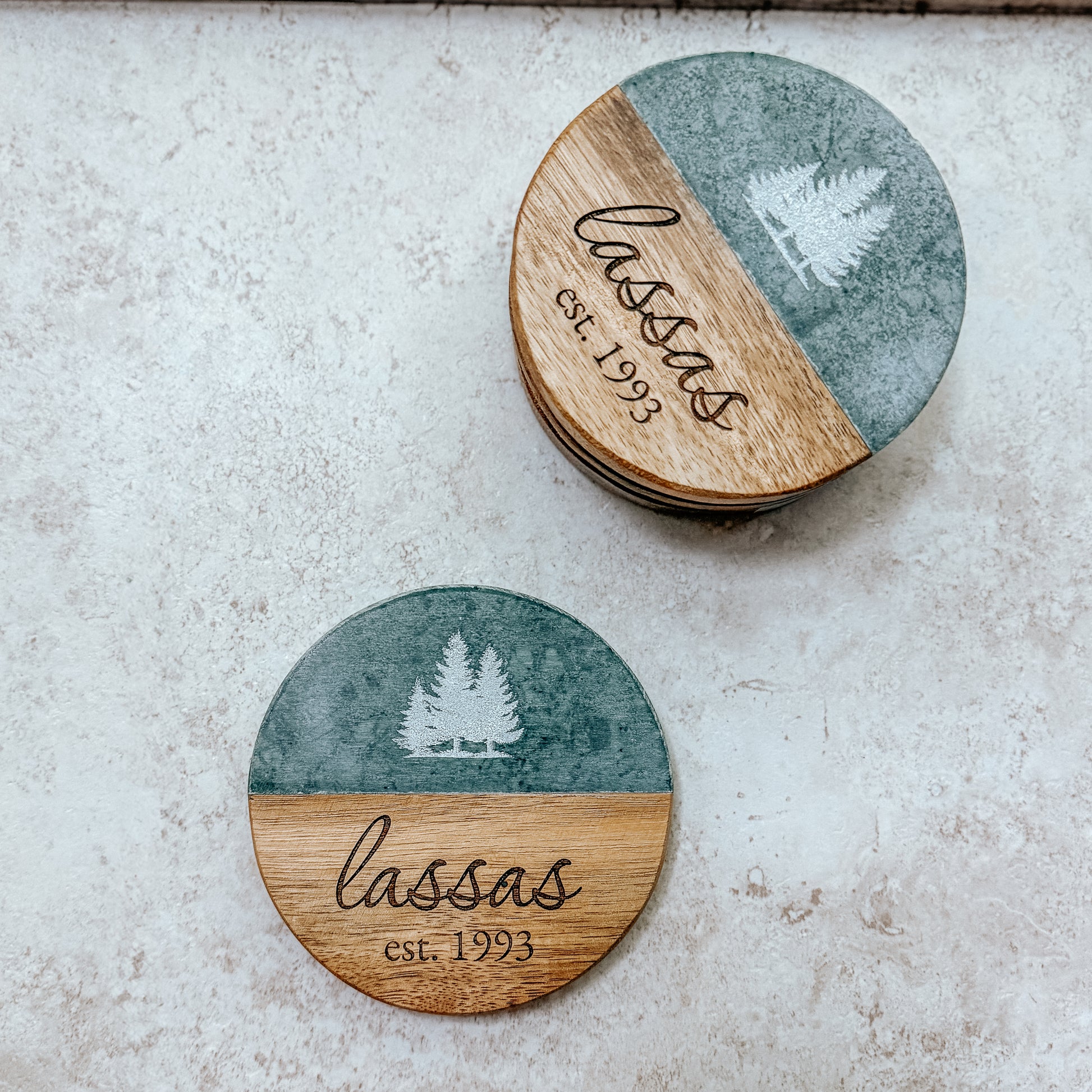 Set of round wooden coasters with green marble-style top, engraved trees, and 'Lassas est. 1993' branding on a light stone background.