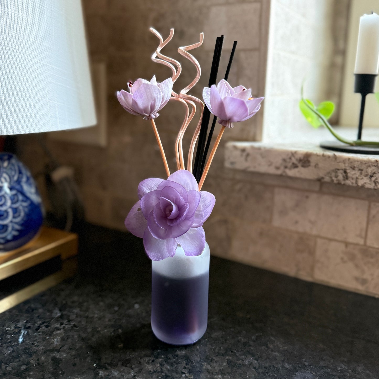 Lilac Bloom reed diffuser with curled floral sticks and black reeds placed near a sunlit window on a dark countertop