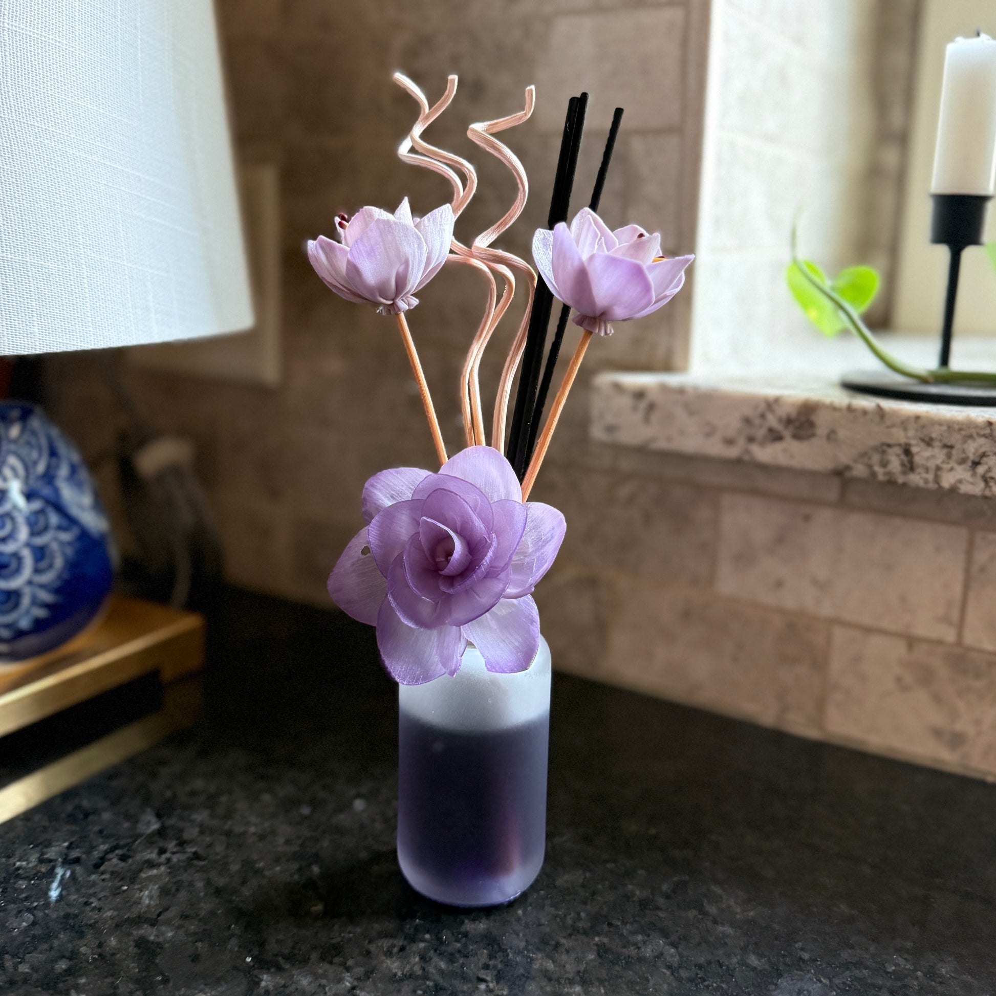Lilac Bloom reed diffuser with curled floral sticks and black reeds placed near a sunlit window on a dark countertop