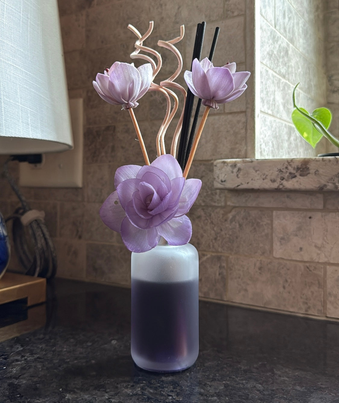 Lilac Bloom reed diffuser with purple floral sticks and black reeds on a kitchen counter beside a window